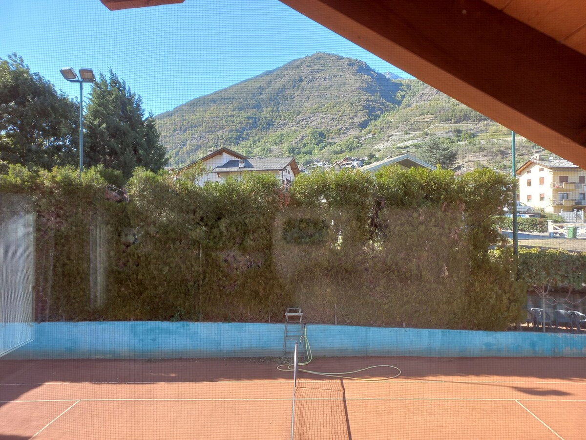 A view from the window reveals a well-maintained tennis court surrounded by green hedges. In the background, a mountain rises, providing a scenic backdrop. The clear blue sky enhances the active atmosphere of the space.