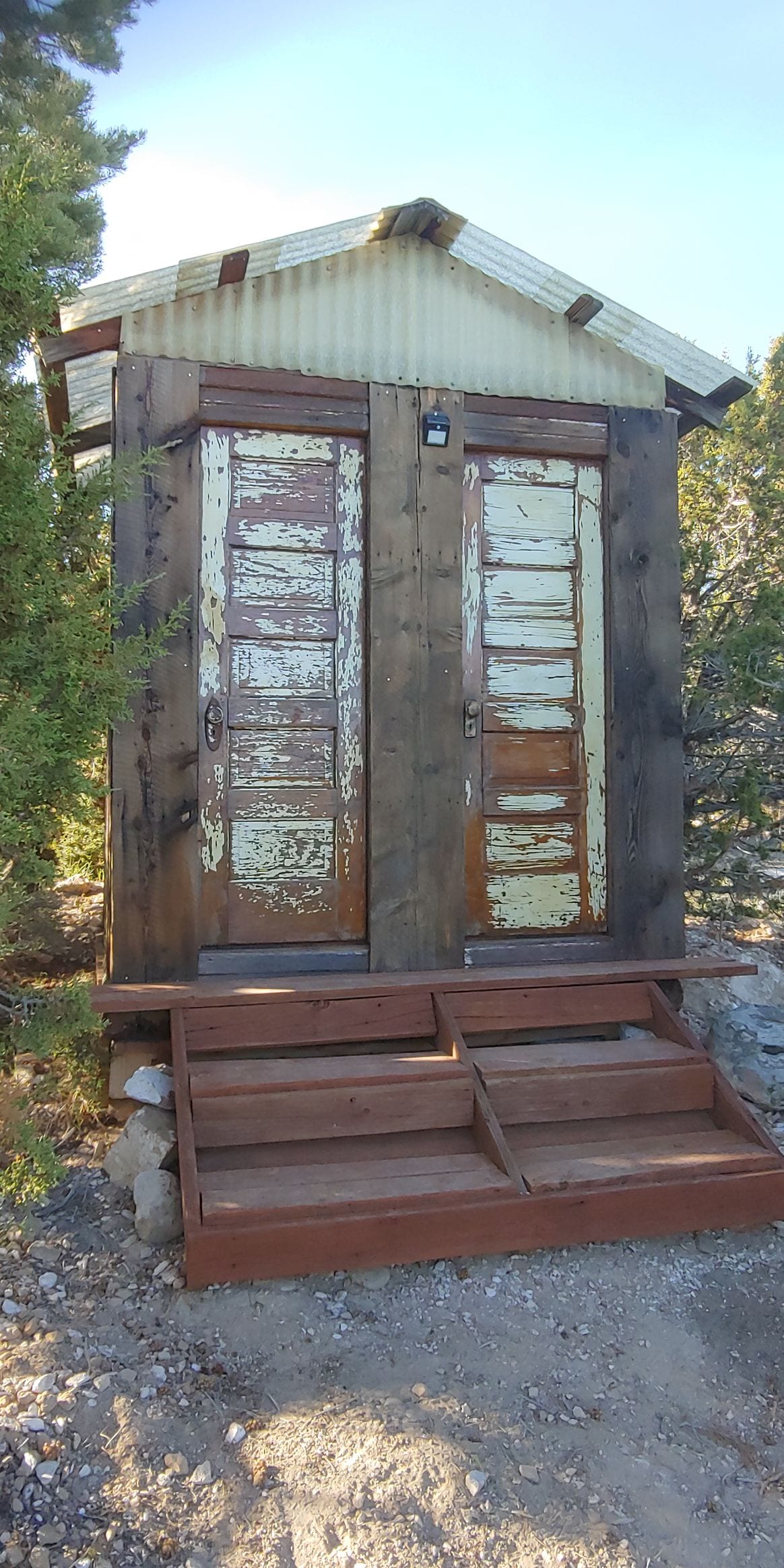 A rustic double outhouse is positioned on a wooden platform, featuring weathered doors with a mix of peeling paint. Steps lead up to the entrance, surrounded by natural foliage, offering privacy. The structure is built amidst the trees, providing a secluded atmosphere for guests.