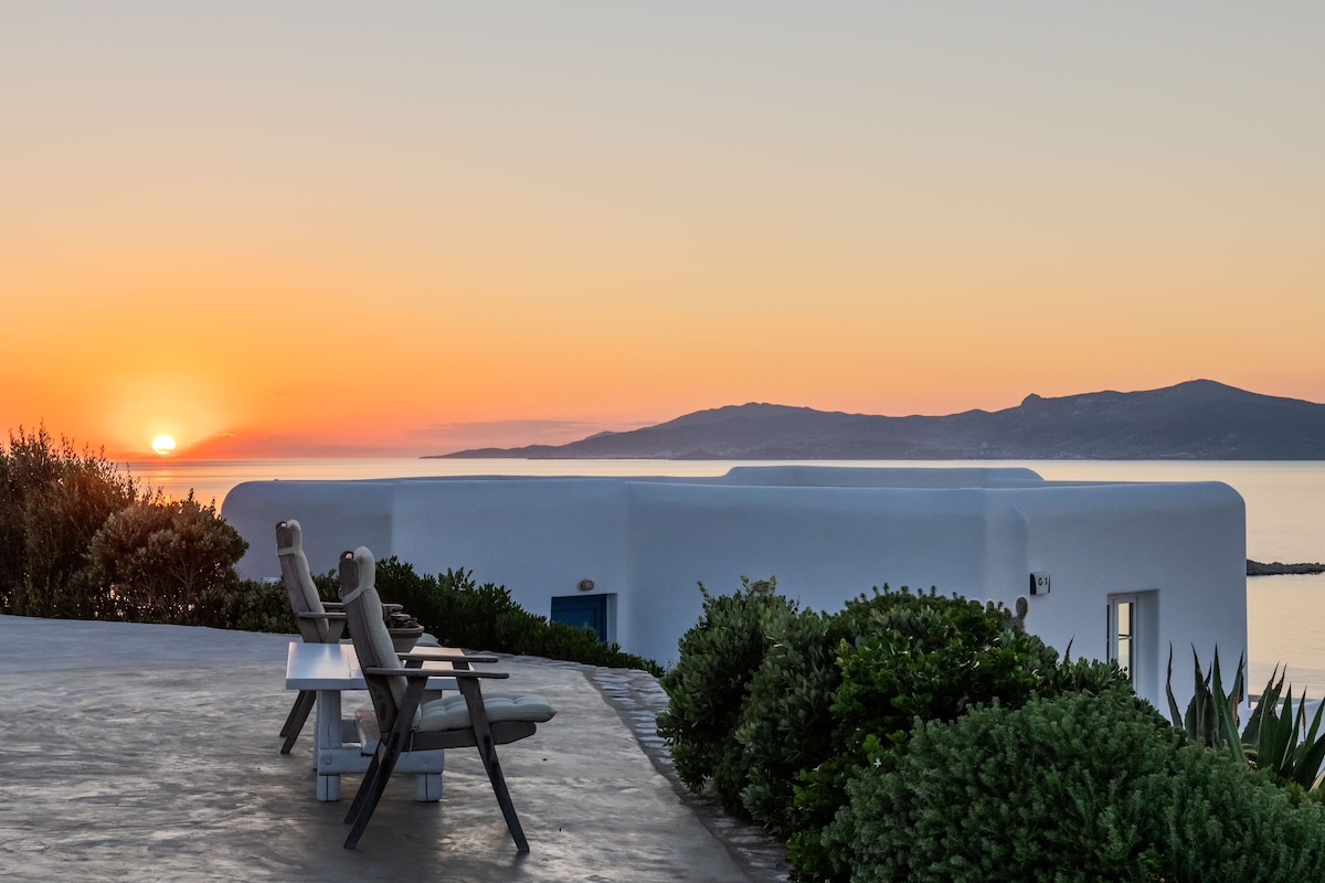An outdoor seating area is shown, featuring two comfortable chairs positioned towards a stunning sunset over the Aegean Sea. The serene landscape includes lush greenery and the silhouette of distant hills, enhancing the tranquil atmosphere of the property.