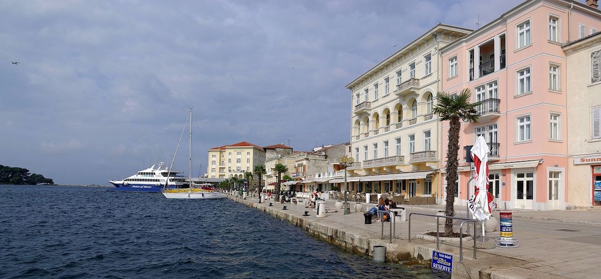 A waterfront promenade stretches along the serene blue water, with elegant buildings lining the street. Palm trees add greenery to the setting, while boats can be seen docked nearby. The scene captures a blend of leisure and urban life amidst cloudy skies.