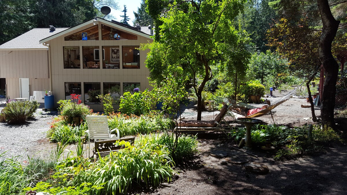 A peaceful garden area is surrounded by lush greenery and flowering plants. A spacious home with large windows overlooks the scene. A hammock is strung between two trees, while outdoor seating offers a place to relax and enjoy the tranquil environment.