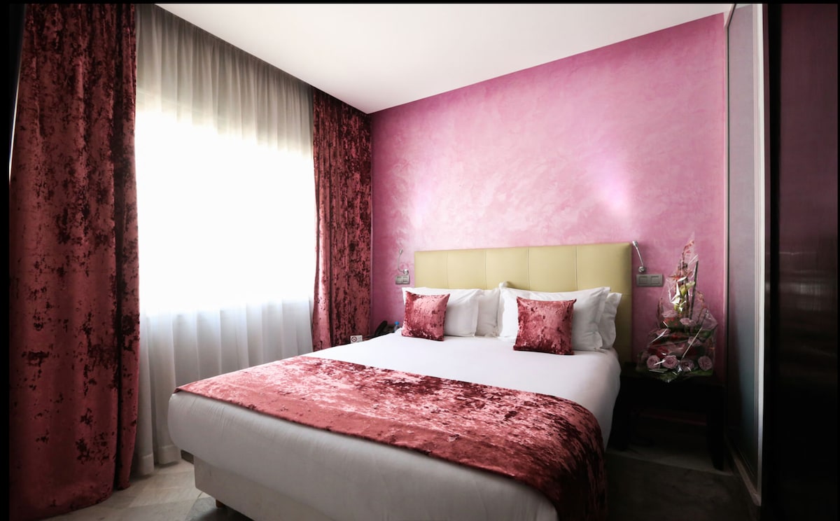 A well-furnished bedroom features a large bed with plush pillows, draped in a soft pink bedspread. The walls display a subtle pink hue, complemented by rich, textured curtains. Natural light filters in through the sheer window treatments, enhancing the room's inviting ambiance.