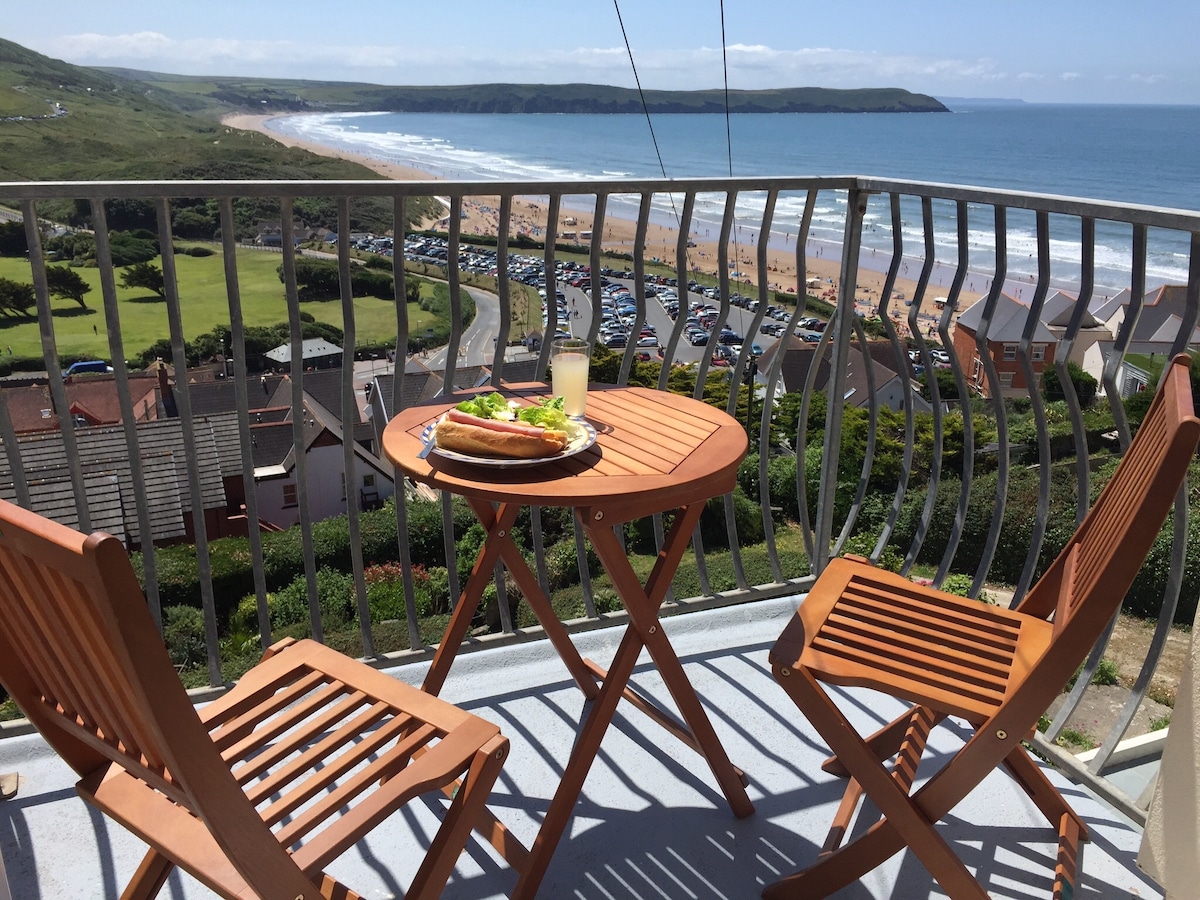 A small balcony features a wooden table set for two, with a plate of food and a drink. The railing offers a view of the sandy beach and surrounding green countryside, with waves visible on the horizon under a clear blue sky.