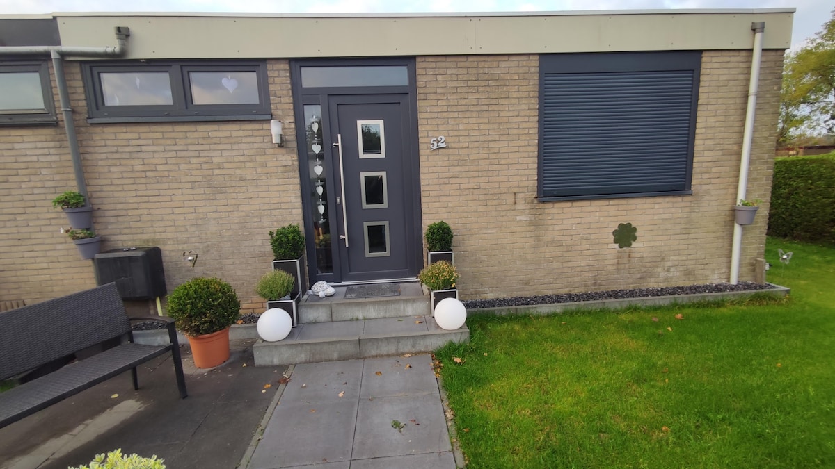 The entrance features a modern door with multiple glass panels and is flanked by potted plants. A few steps lead up to the door, with a small patio area partially covered in grass. The exterior wall is finished in a light brick pattern.