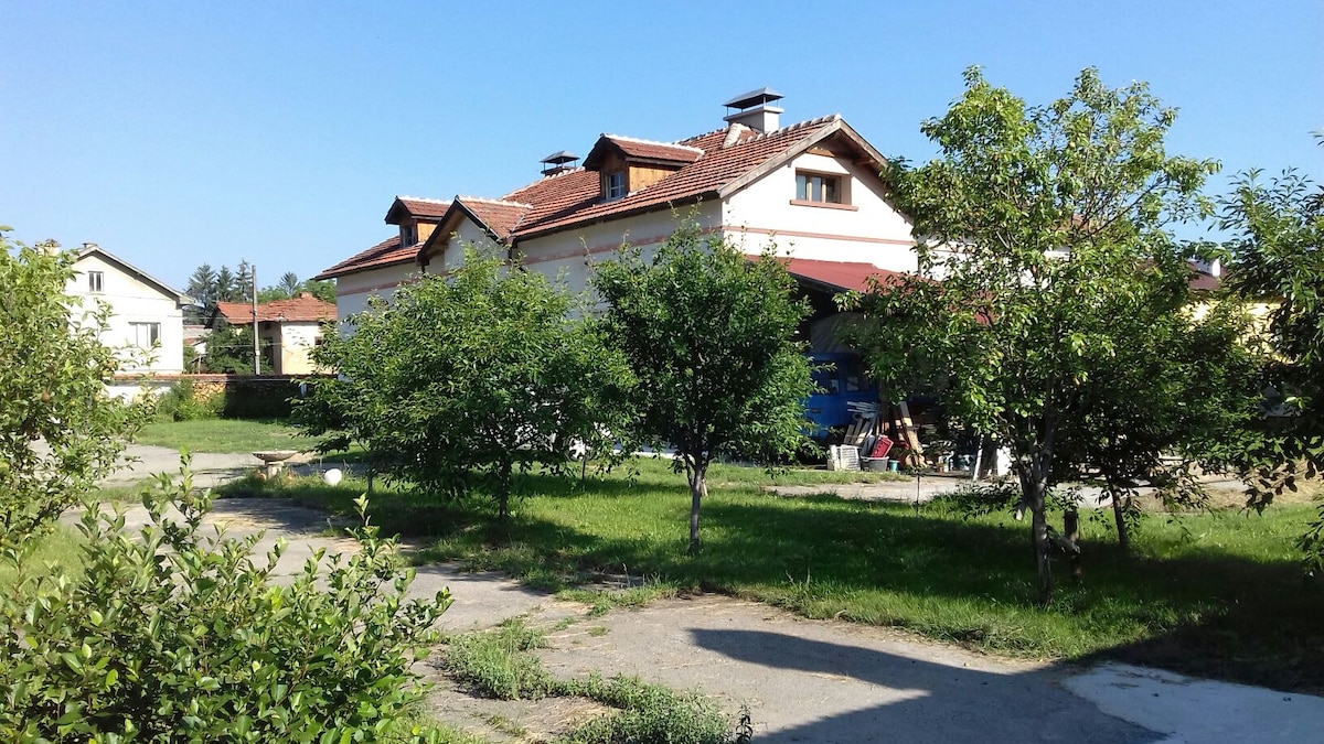 A spacious outdoor area is surrounded by greenery, featuring several trees and a well-maintained lawn. A large villa with a tiled roof is visible in the background, showcasing a welcoming entrance covered by a porch. The surrounding environment offers a serene countryside atmosphere.