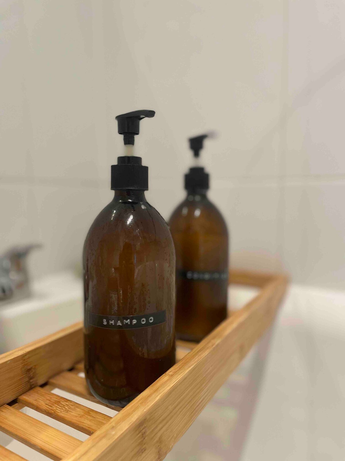 Two amber glass bottles labeled as shampoo are positioned on a bamboo bath caddy. The bottles feature black pumps and are set against a light-tiled background, highlighting a minimalist and functional design in the bathroom.