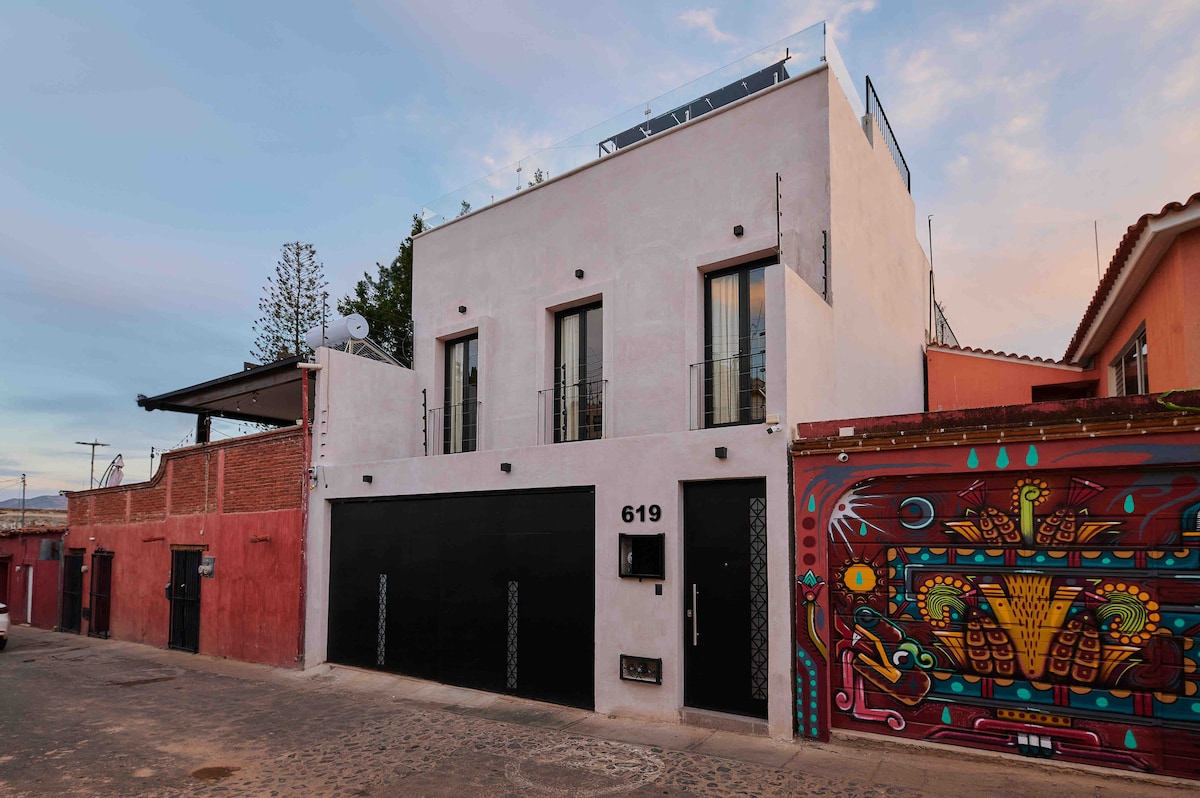 A modern boutique house features a minimalist design with smooth, white walls and large windows, providing a sense of openness. A vibrant mural in multiple colors decorates the adjacent wall, adding a local artistic touch to the surroundings.