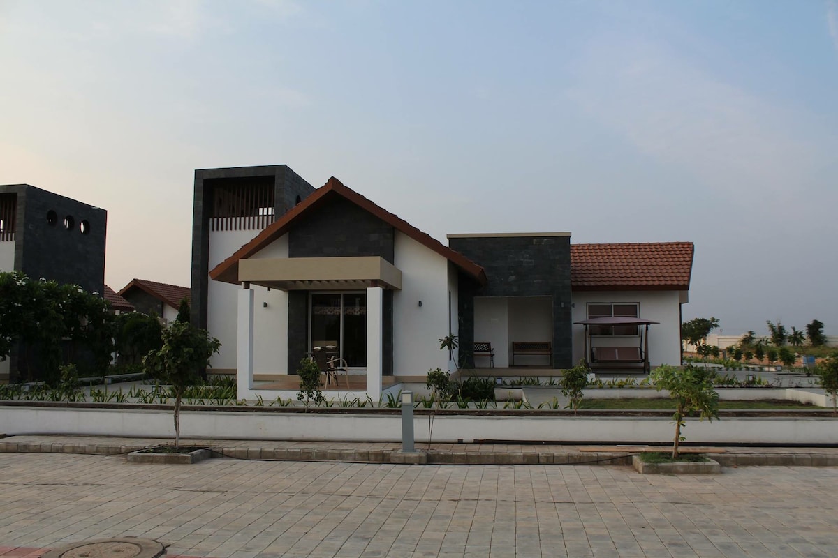 A modern farmhouse is displayed, featuring a combination of stone and white walls. The roof has a warm terracotta hue. Large windows allow natural light to enter, while neatly maintained greenery surrounds the property, enhancing its serene setting.