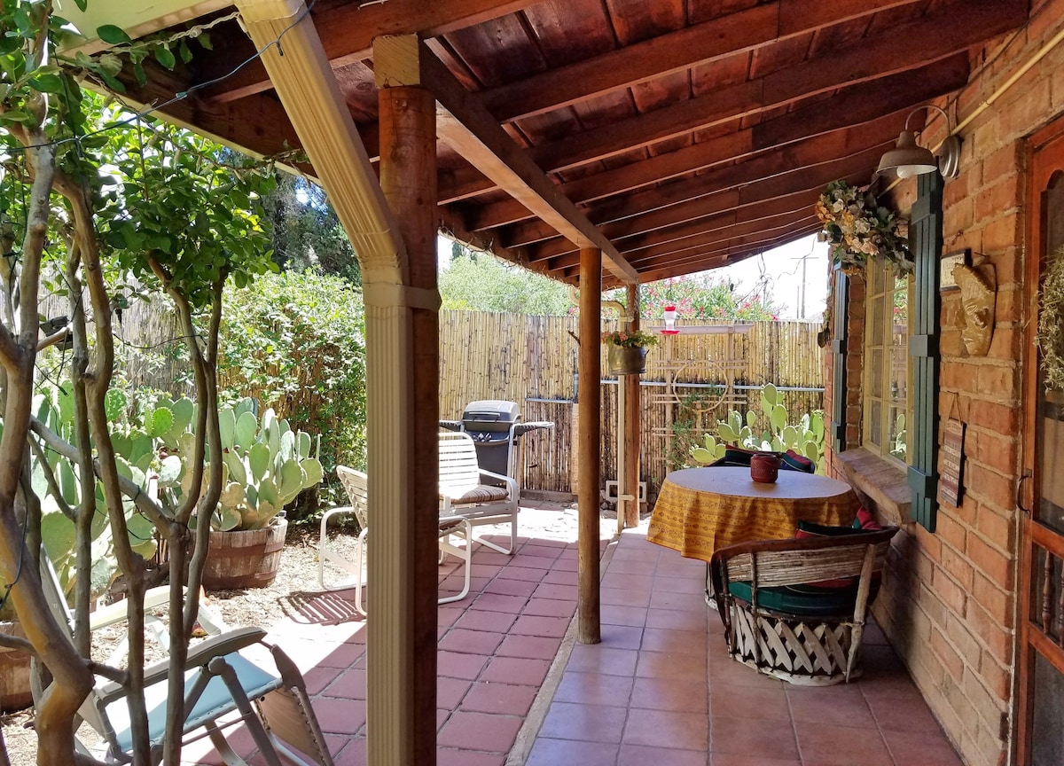 A shaded patio area features a round table with a vibrant tablecloth, surrounded by comfortable seating. Lush greenery, including cacti and flowering plants, is visible in the garden, enhancing the natural setting. A barbecue grill is positioned nearby for outdoor cooking.