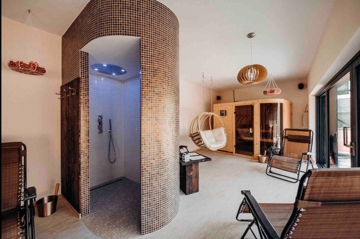 A private sauna area features a circular tiled shower and a hanging chair, providing a space for relaxation. Comfortable lounge chairs are arranged strategically, with large windows offering natural light and views of the surrounding area.