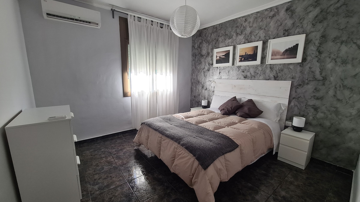 A spacious bedroom features a queen bed with soft bedding, complemented by decorative pillows. Natural light filters through sheer curtains, illuminating the room. Adjacent white nightstands and a dresser provide practical storage. A decorative pendant light hangs above, enhancing the serene atmosphere.