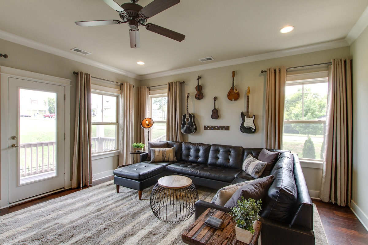 A modern living room features a dark sectional sofa positioned around a decorative coffee table. Natural light fills the space through large windows, complemented by gentle curtains. A rustic side table and wall-mounted guitars add character to the inviting atmosphere.