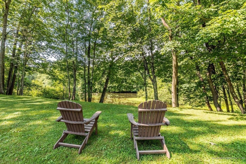 Two wooden adirondack chairs are positioned on a grassy area, facing the tranquil Mousam River. Lush trees provide a natural canopy, offering shade and enhancing the serene atmosphere of the surrounding garden.