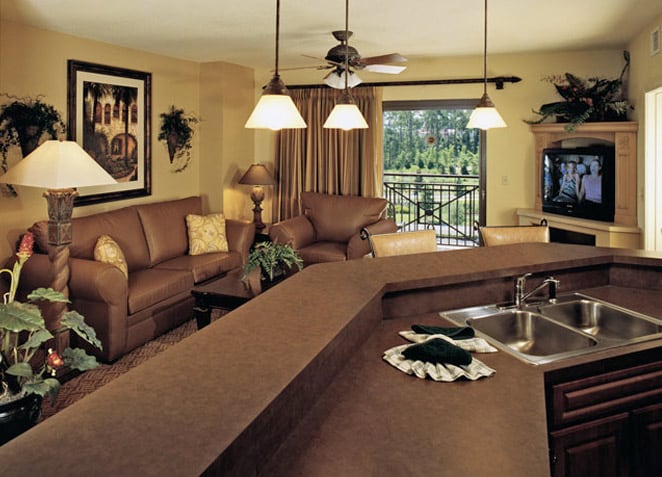 A spacious living area features a comfortable seating arrangement with brown leather sofas and decorative pillows. Natural light filters through large windows, illuminating the space. An entertainment unit displays a television, and a kitchen bar is visible with a sink and modern countertop.