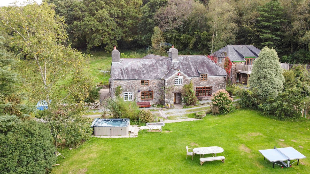 The exterior of the historic stone farmhouse is visible, nestled amidst lush greenery. A spacious garden features a wood-fired hot tub, outdoor dining area with a circular table, and lounge chairs. The surrounding trees offer privacy, while a rustic outbuilding can be seen in the background.