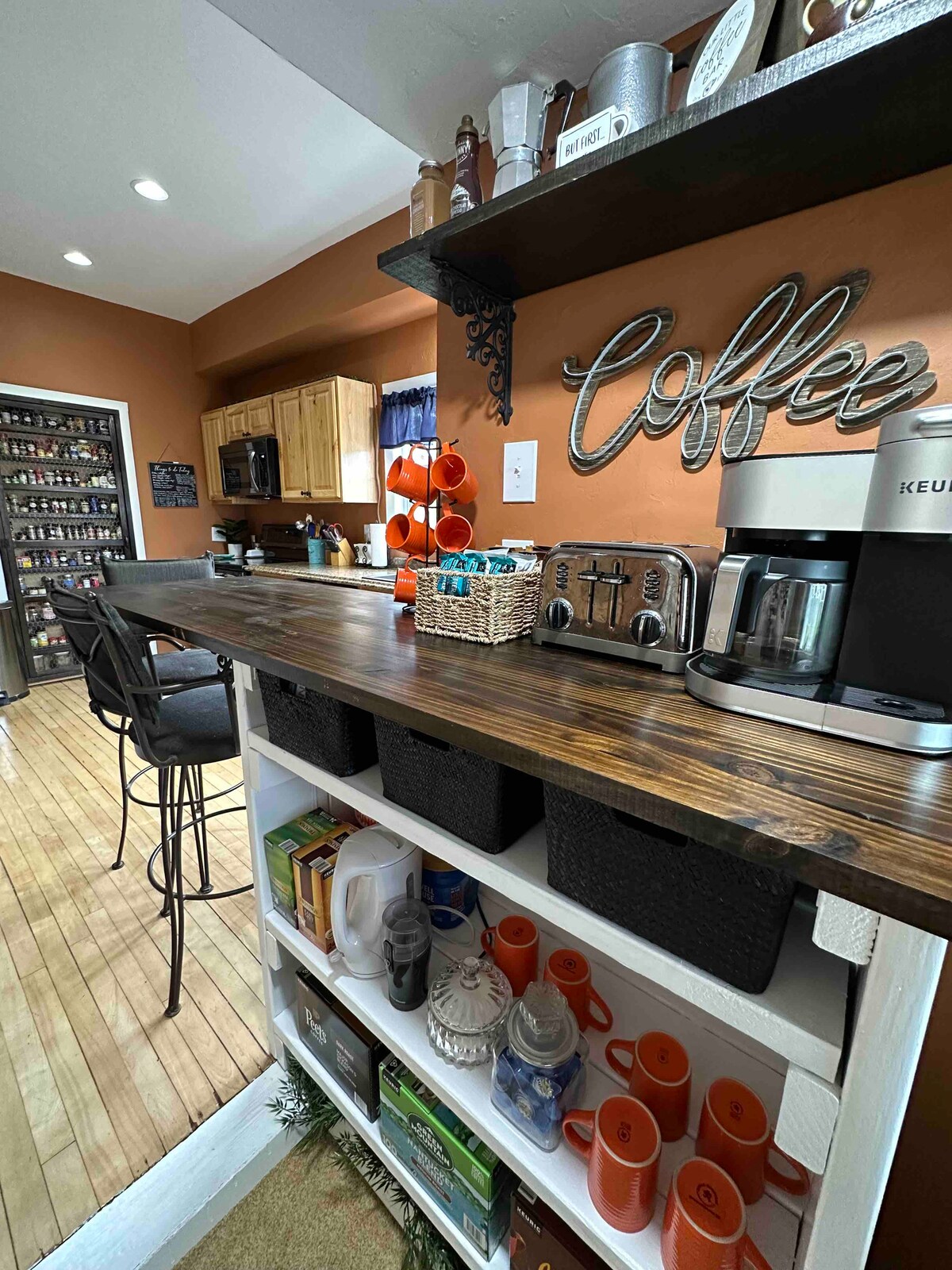The kitchen features warm, earthy tones with wooden cabinetry and a coffee bar. A coffee maker, toaster, and decorated shelving are visible, alongside organized baskets. Bar stools line the counter, enhancing the space's functionality and inviting atmosphere.