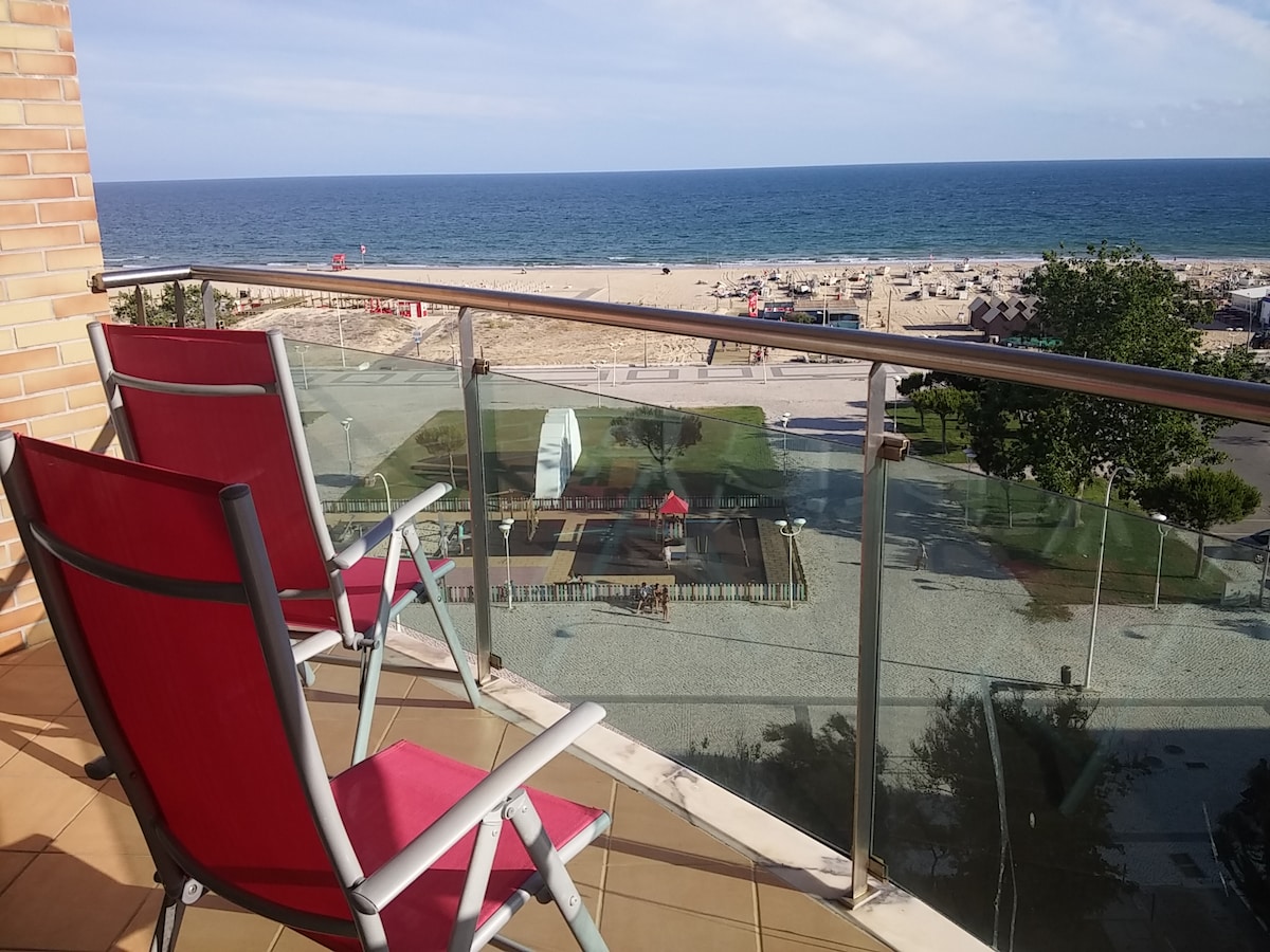 A balcony with two red chairs provides a view of the sandy beach and ocean. The space is enclosed by a glass railing, allowing unobstructed visuals of the horizon. Below, a playground and beach area are visible, with sun loungers arranged along the shore.