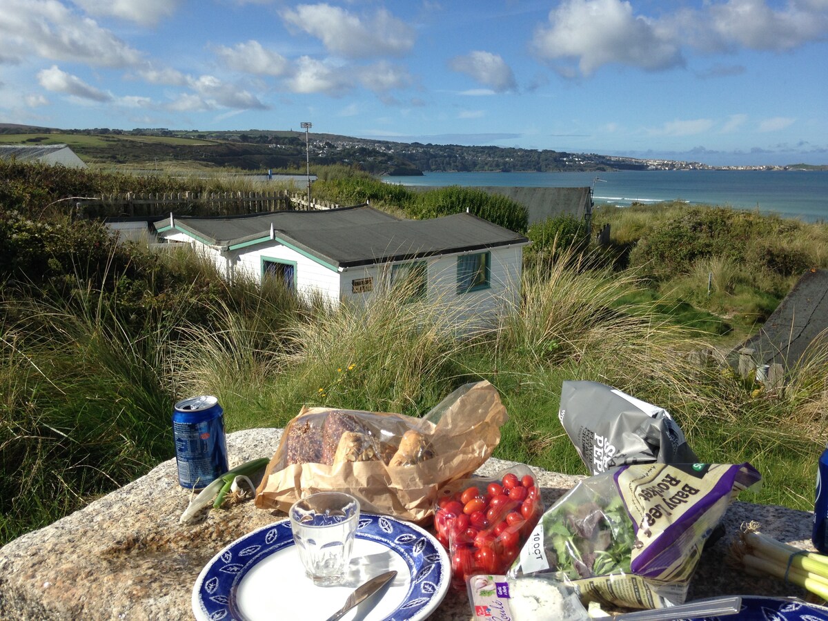 A picnic area is depicted with a stone table featuring a glass, plates, and assorted food items. The surrounding grass and dunes frame a distant view of the coastline and a nearby chalet, under a partly cloudy sky. The tranquil sea is visible in the background.