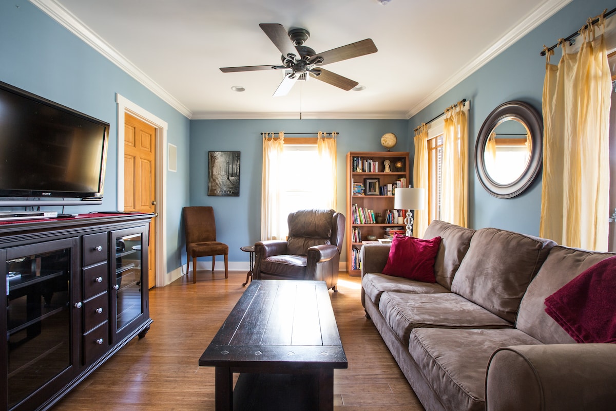 The living room features a comfortable, spacious couch with accent pillows. A wooden coffee table is situated in front of the couch, complementing a sleek television stand. Natural light enters through multiple windows adorned with light curtains, illuminating the room's warm colors.