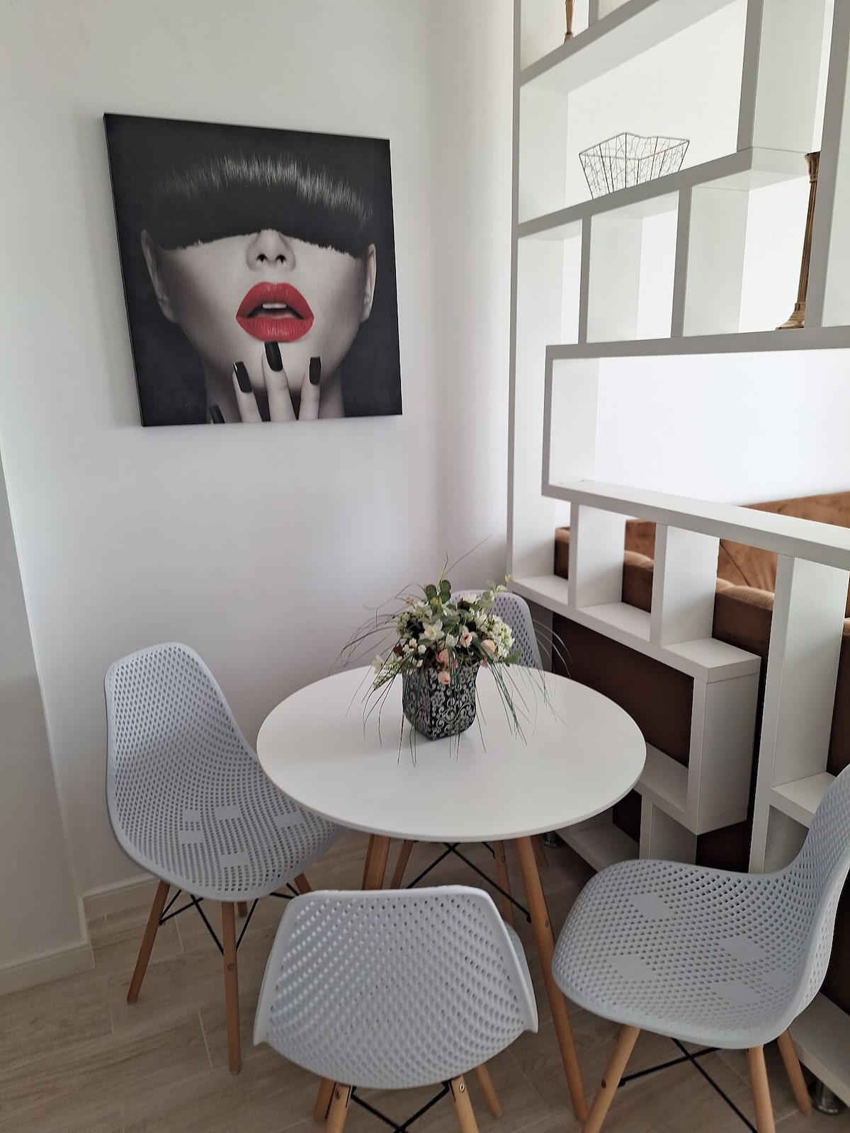 A small dining area is presented, featuring a round white table surrounded by three light grey chairs with wooden legs. A decorative vase filled with flowers sits at the center. A piece of art hangs on the wall, adding visual interest to the space.