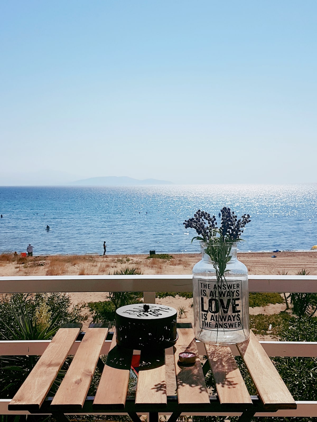A serene balcony view showcases the sparkling sea and sandy beach, with distant waves gently lapping at the shore. A wooden table is positioned in the foreground, adorned with a decorative jar of lavender and a compact black object, inviting relaxation and connection to the coastal scenery.