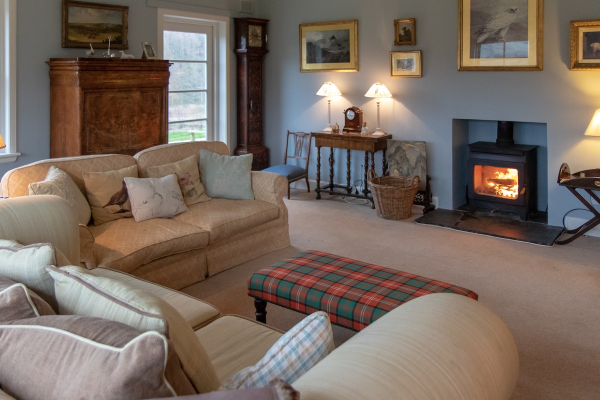 A spacious sitting room features a cozy wood-burning stove, surrounded by comfortable seating and a patterned ottoman. Natural light enters through large windows, highlighting framed artwork on the walls. Soft lighting from table lamps creates a warm and inviting ambiance.