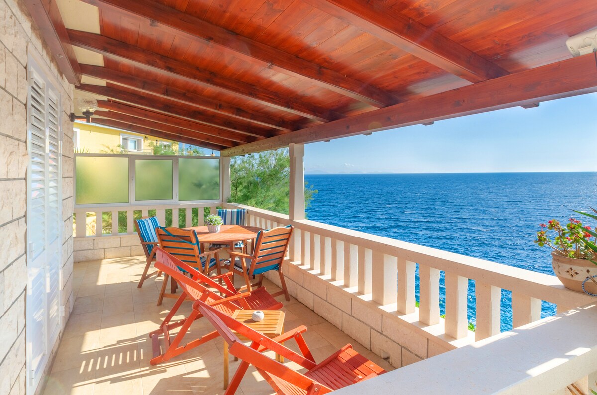 A covered terrace features wooden beams and a stone railing overlooking the sea. It includes a round wooden table surrounded by four chairs with bright blue and red slats. Potted plants add greenery to the space, enhancing the coastal view.