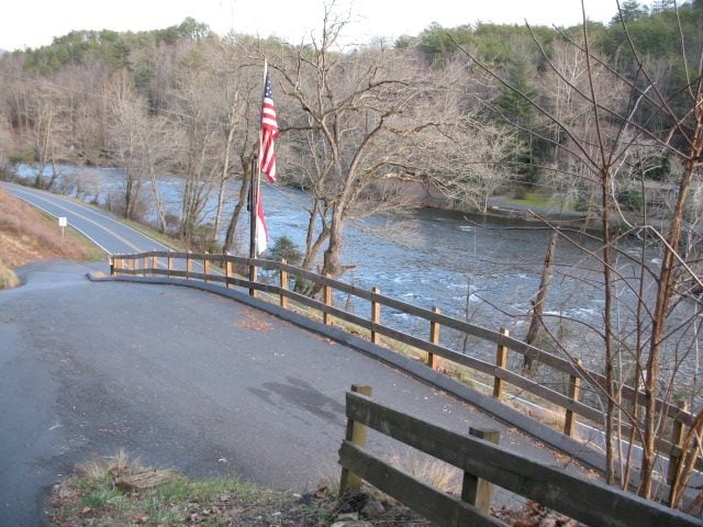 A winding road leads down towards the Tuckaseegee River, with a wooden fence lining one side. An American flag is displayed prominently, while trees with bare branches stand along the riverbank. The water flows gently, reflecting the surrounding landscape.