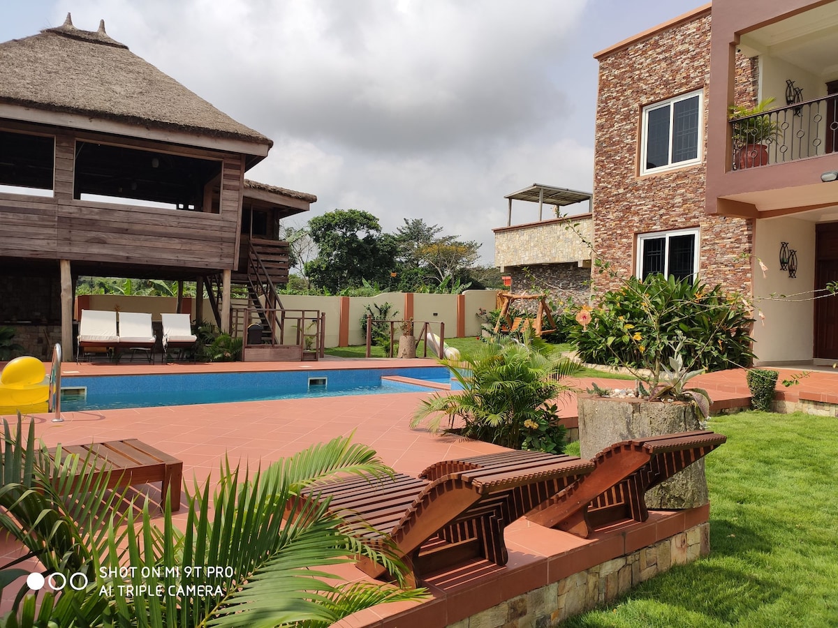 An outdoor area is featured with a swimming pool surrounded by lush greenery. Two wooden lounge chairs are positioned near the pool, and a summer hut is visible in the background. The setting is tranquil, providing ample space for relaxation and outdoor activities.