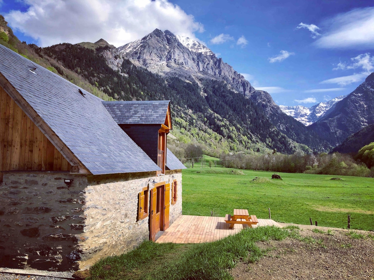 A rustic stone building is set against a backdrop of mountains, with green fields surrounding it. A wooden deck is visible, featuring a picnic table that invites outdoor gatherings. The sky is bright, highlighting the natural beauty of the landscape.