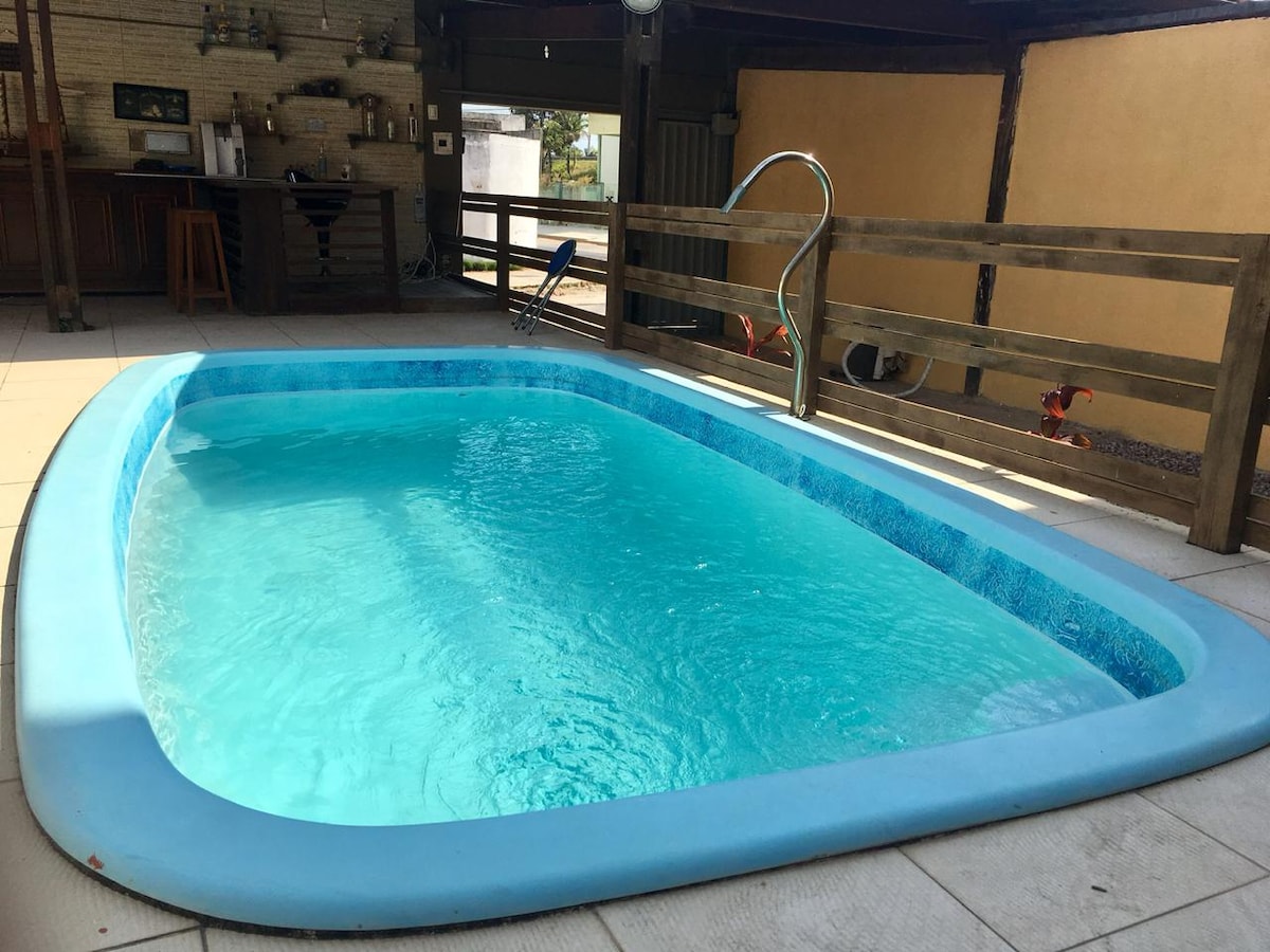 A private swimming pool is featured within a shaded outdoor area. The water is clear and inviting, surrounded by tiled flooring. A wooden railing lines one side of the pool, and a small ladder provides access for entry. Bar stools and a bar area are visible in the background.