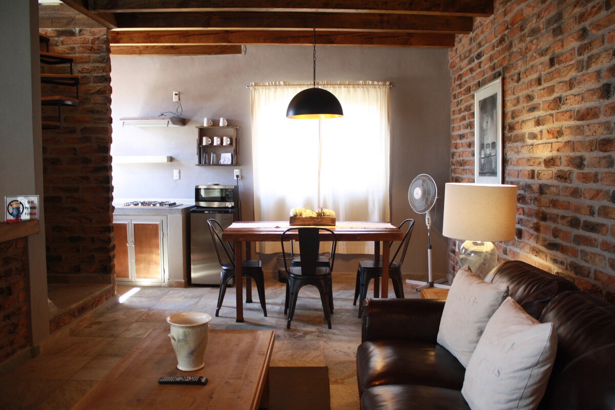 An inviting loft space features a rustic brick wall, a wooden dining table with metal chairs, and a well-equipped kitchen area. Natural light fills the room through a large window, complemented by a stylish pendant light. A comfortable sofa adds to the cozy atmosphere.