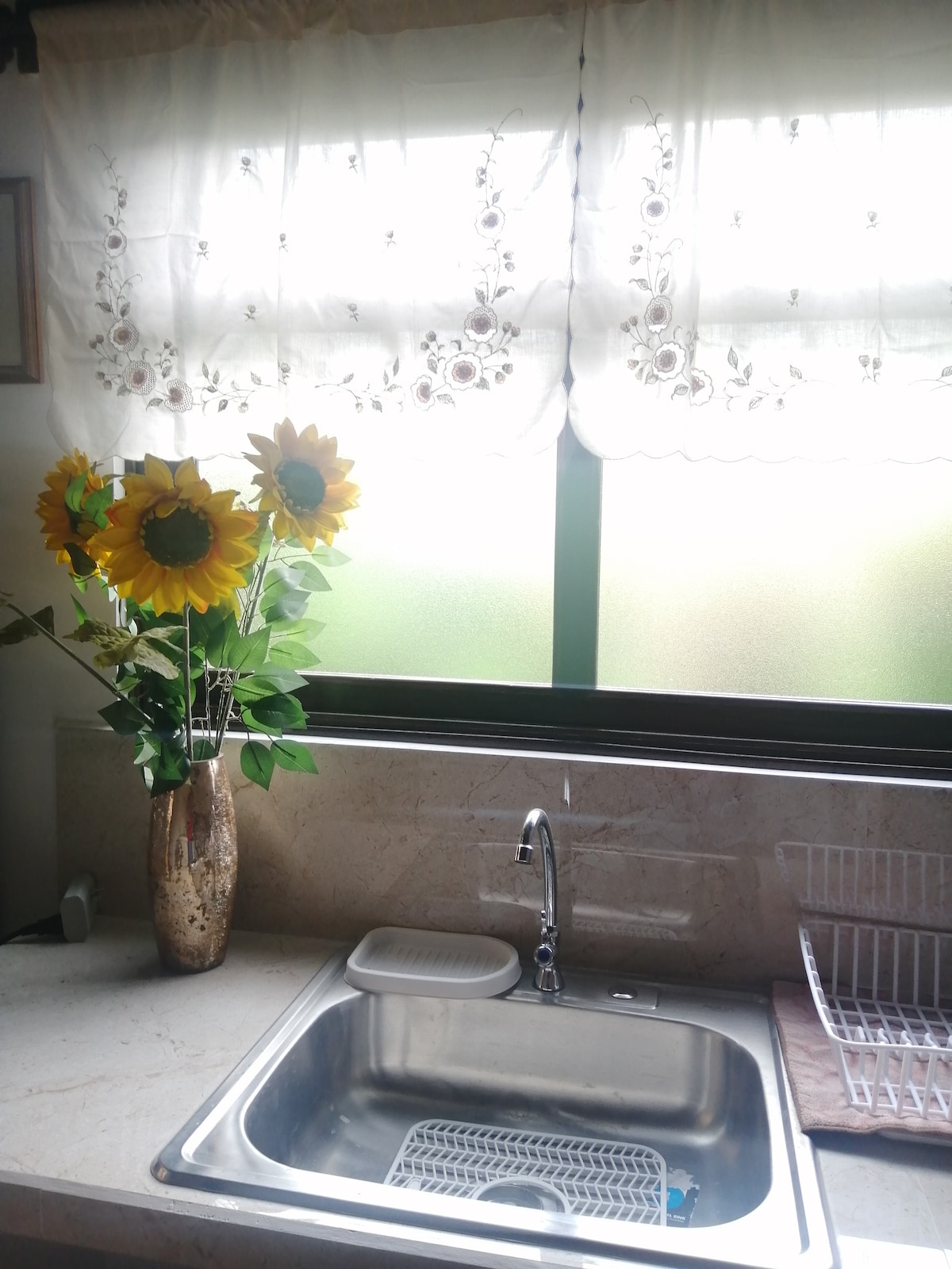 A kitchen sink is placed on a marble countertop, accompanied by a dish drying rack. In front of the window, decorated with lace curtains, a vase filled with fresh sunflowers adds a touch of color and brightness to the space.