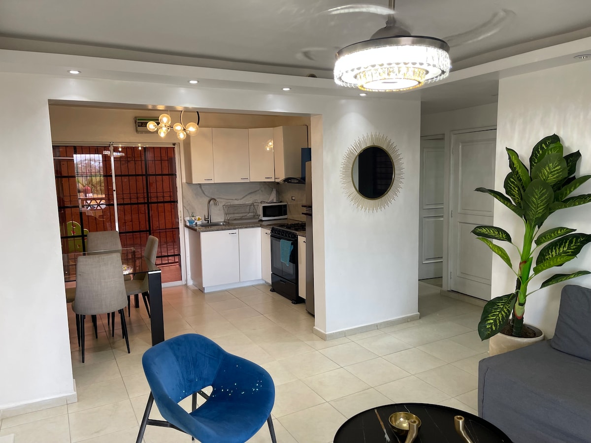 A modern living area is shown with a spacious layout, featuring a gray sofa and blue accent chair. The kitchen area, visible in the background, includes white cabinetry and a dining table with chairs. A circular mirror and indoor plant add a touch of decor.
