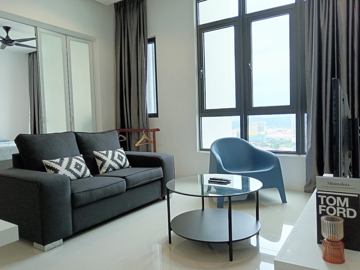 A modern living area features a dark sofa with geometric cushions, complemented by a round glass coffee table. Large windows allow natural light to fill the space while offering scenic views. A minimalistic side table displays a decorative item, adding a touch of sophistication.