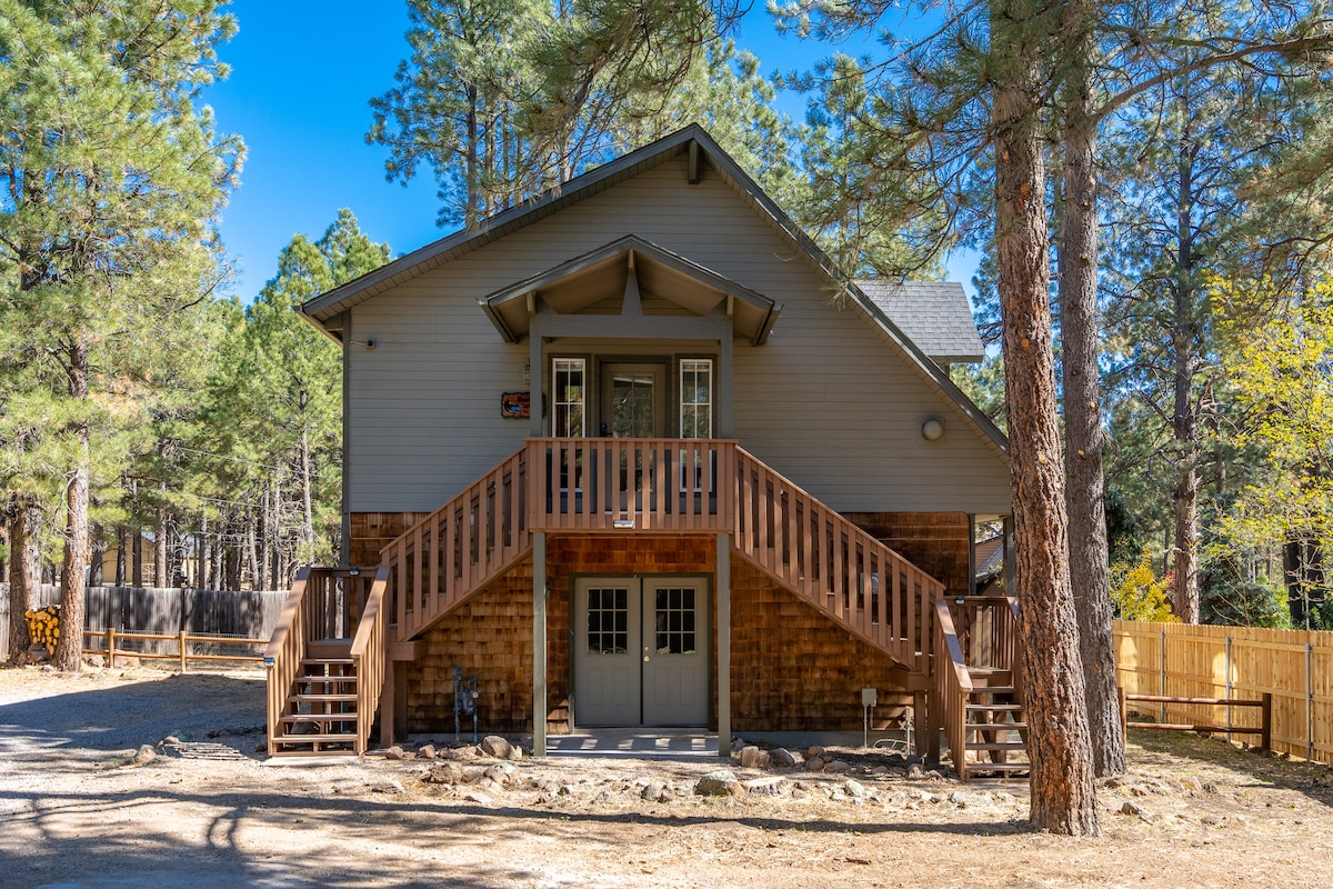 The exterior of a two-story cottage is featured, showcasing wooden steps leading to a double door entrance. The structure is surrounded by tall pine trees and gravel pathways, with a fenced yard visible in the background.