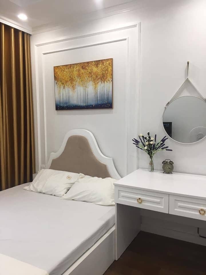 A neatly arranged bedroom features a spacious bed with light bedding against a backdrop of golden curtain panels. A decorative painting of trees adorns the wall above the bed. A mirror and a small vase with flowers are placed on a white desk, providing a functional and stylish touch.