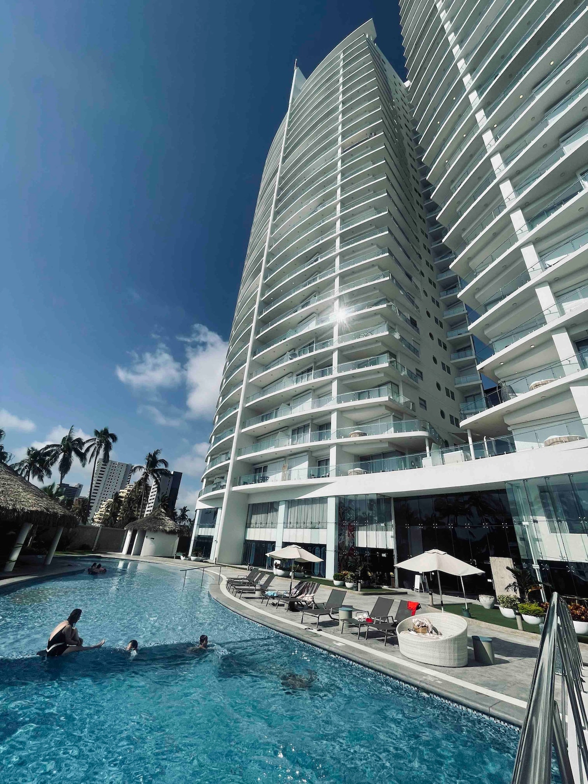 A tall, modern building is depicted with multiple balconies, reflecting sunlight against a clear blue sky. A spacious pool is shown in the foreground, surrounded by lounge chairs and umbrellas, while guests enjoy the water. Lush palm trees are visible nearby.