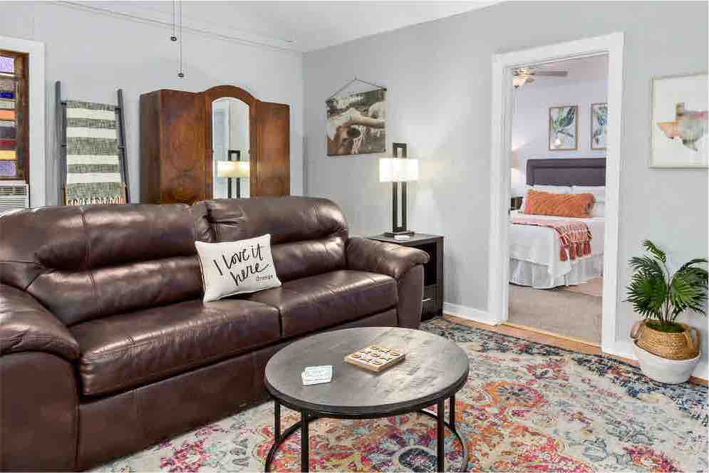 An inviting living area features a comfortable brown leather sofa, complemented by a round coffee table on a colorful area rug. A well-lit corner showcases a lamp and potted plant, while a doorway leads to a cozy bedroom in the background.