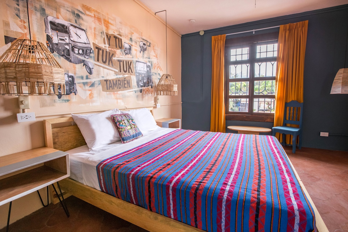 A cozy bedroom is presented with a colorful striped blanket draped over a spacious bed. Natural light filters through a large window, flanked by warm yellow curtains. Unique woven light fixtures add texture, complemented by a vibrant wall mural featuring cultural elements.