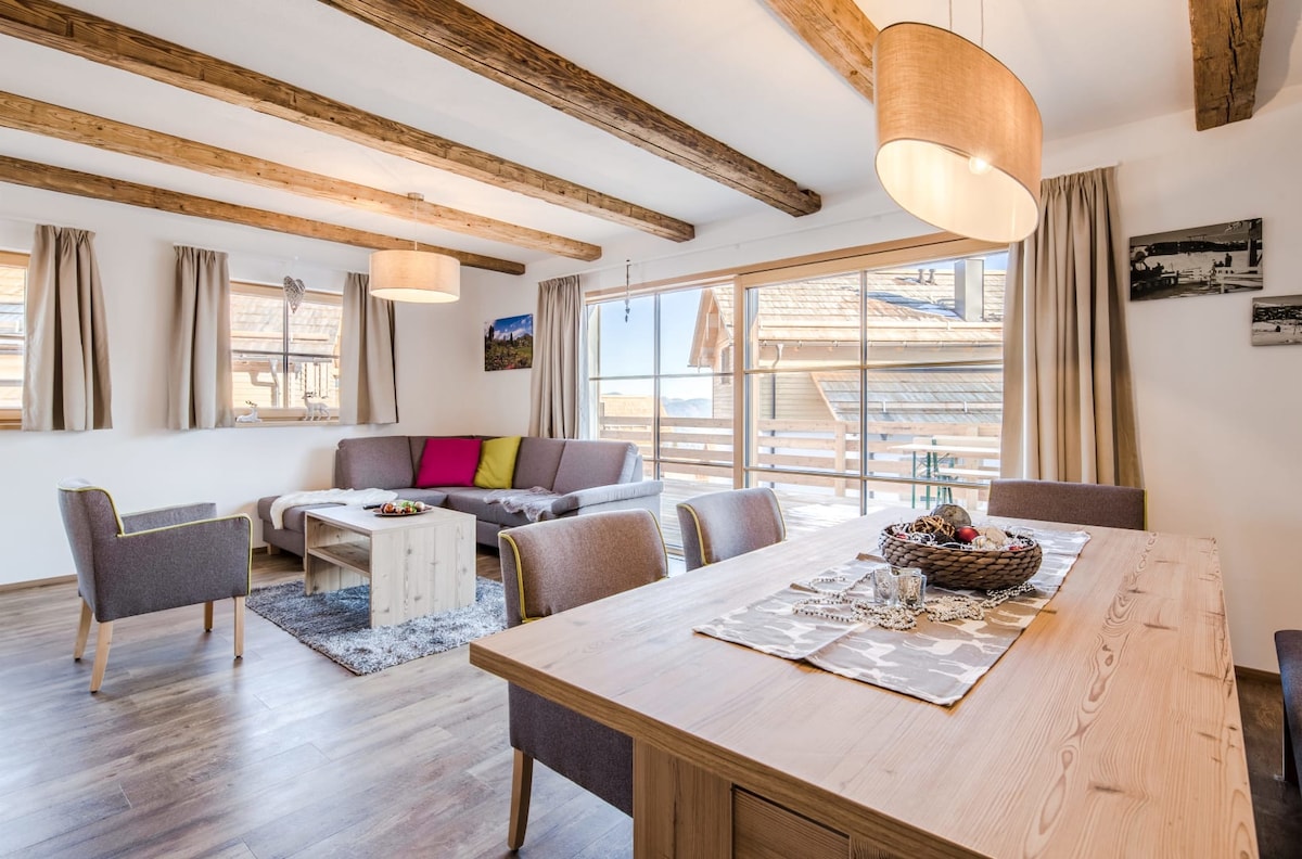 An open-plan living and dining area features wooden beams across the ceiling, enhancing the room's spaciousness. A large dining table sits beside a cozy seating arrangement, which includes a sofa and armchairs. Natural light floods in through expansive windows, offering views of the exterior.