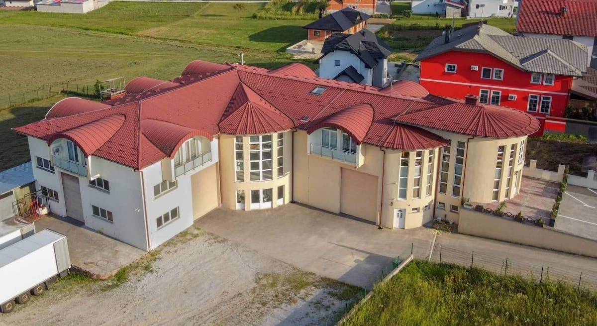 An expansive luxury apartment complex is shown, featuring a distinctive curved roof and large glass windows. The building's facade exhibits a mix of pale and cream colors, surrounded by open green spaces and neighboring residences. Ample parking space is visible in front of the structure.