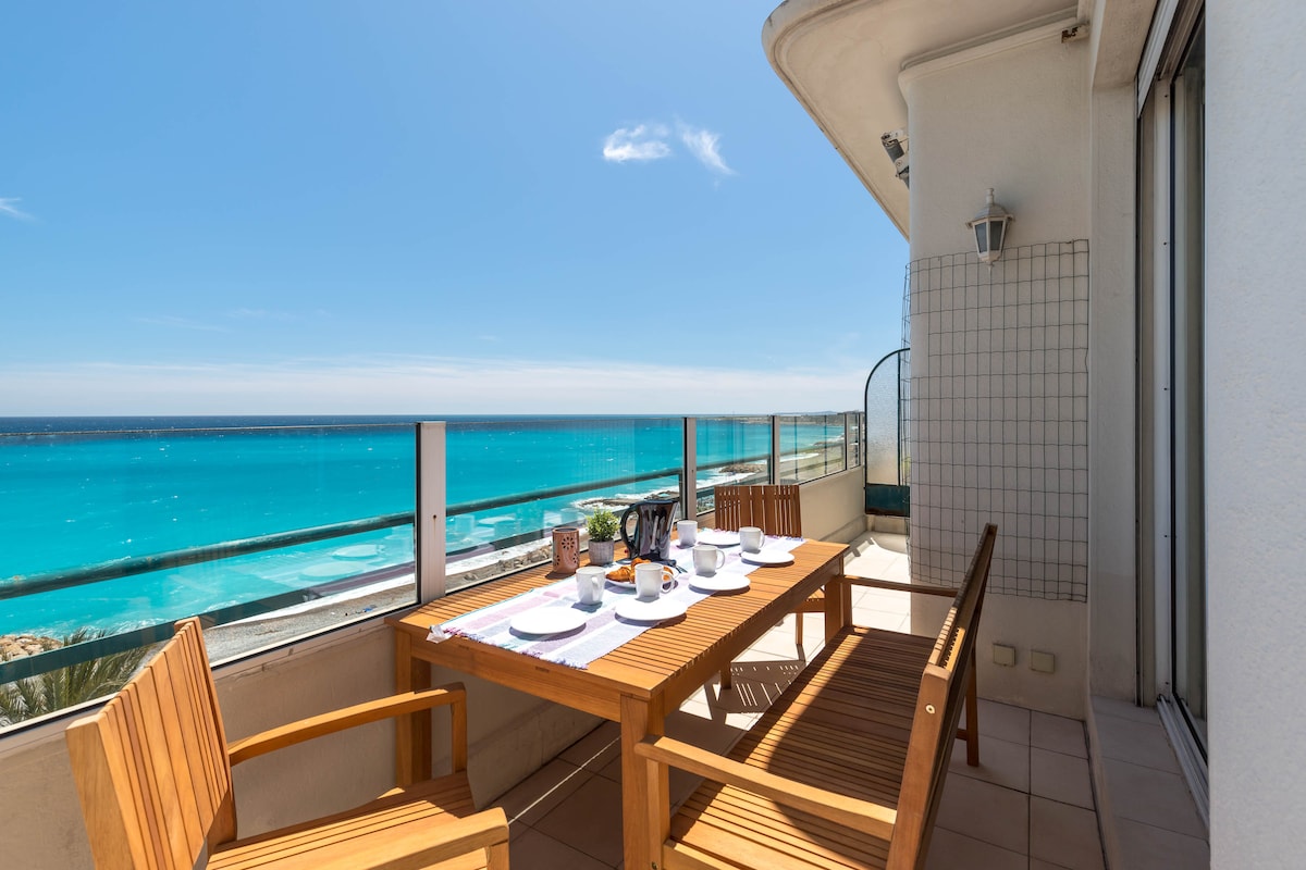 A terrace with a wooden dining table is positioned against a panoramic view of the sea. The table is set for a meal with plates, cups, and a small centerpiece. Clear skies and vibrant turquoise waters create a bright and inviting atmosphere.