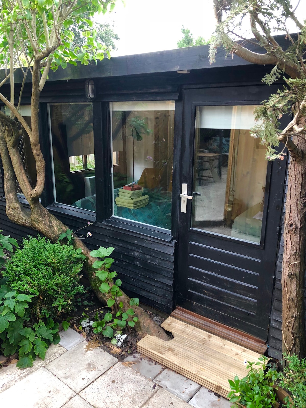 The entrance of the garden house is framed by lush greenery and trees, showcasing a wooden door with a large window. The exterior is painted black, highlighting the home’s charming design against the natural surroundings.