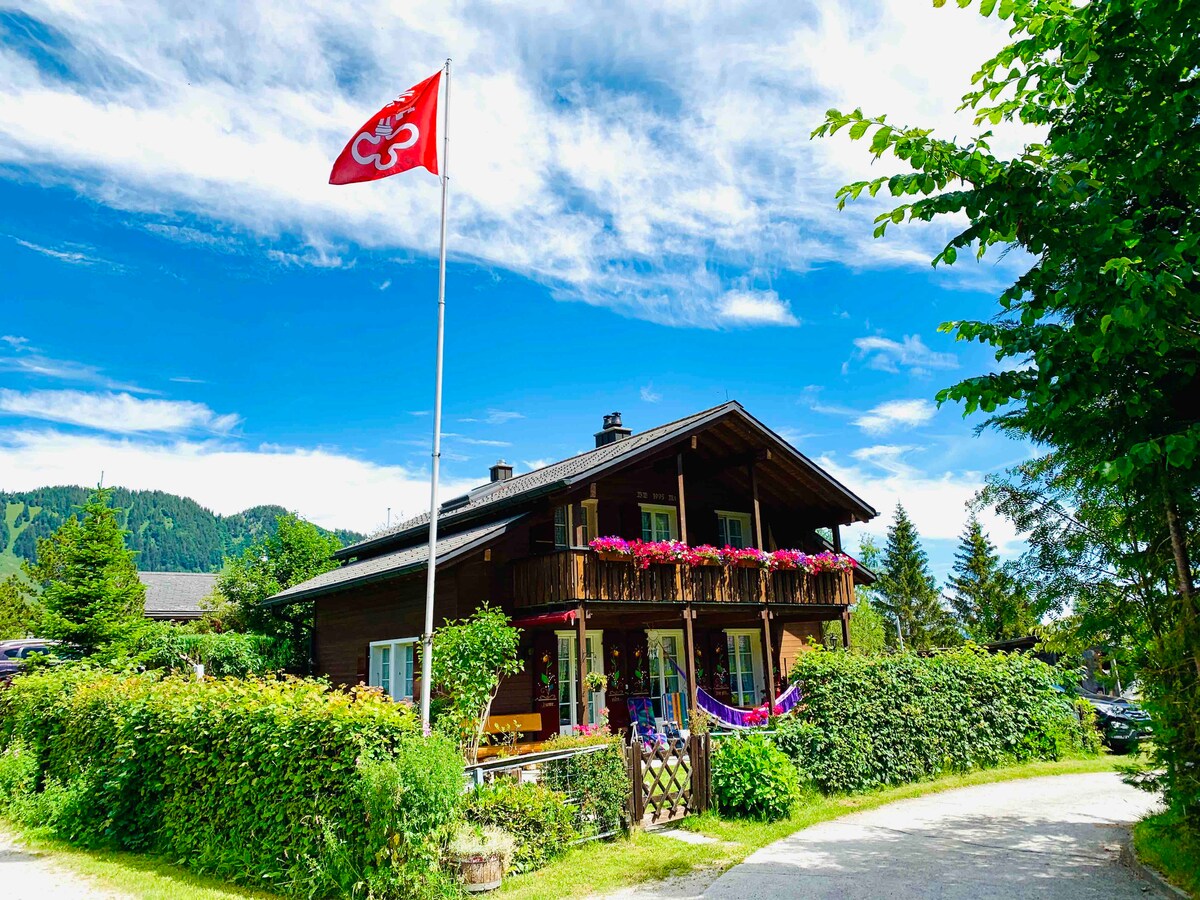 A chalet-style building is surrounded by lush greenery and colorful flower boxes, displaying vibrant blooms on the balcony. A flag proudly features the Swiss emblem against a clear blue sky, while distant mountains are visible in the background, creating a serene alpine atmosphere.