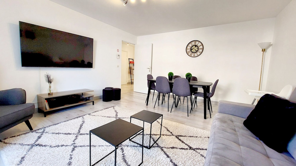 A bright living area features a large flat-screen TV mounted on the wall, accompanied by a gray sofa and a matching armchair. A low coffee table is situated on a textured rug, with a dining table and chairs positioned nearby, enhancing the open layout.