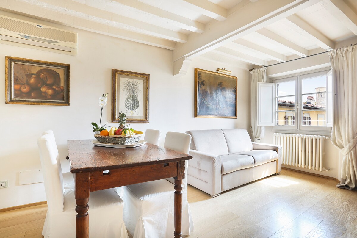 A dining area features a rustic wooden table surrounded by four white chairs. A fruit bowl is displayed at the center of the table. A light-colored sofa sits against the wall, with large windows inviting natural light. Two framed artworks are hung on the walls.