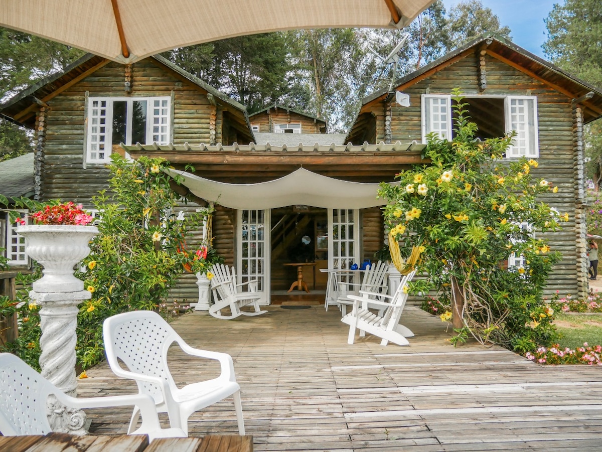 The wooden cabin, constructed from log materials, features a spacious deck adorned with white rocking chairs. Surrounding greenery and blooming flowers enhance the inviting outdoor space. The cabin's large windows provide a view of the interior, creating a welcoming entrance.