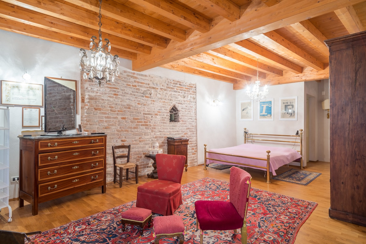 A spacious room features exposed wooden beams and a combination of brick and white walls. An antique dresser and a red velvet chair are positioned near the elegant bed. A decorative rug covers the wooden floor, and light is provided by two chandeliers.