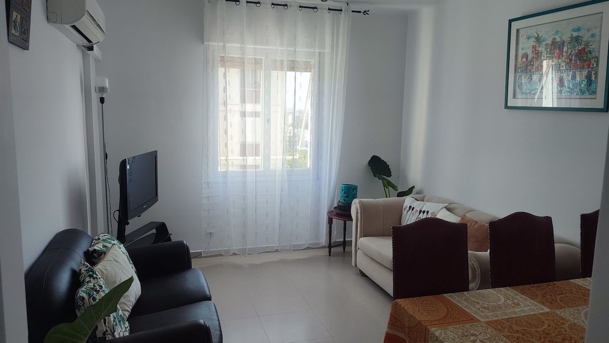 An airy living room features two sofas, one in black leather and another in cream fabric, arranged around a small wooden table. Natural light enters through a large window, accentuated by sheer curtains. A television sits on a wall-mounted bracket, and a potted plant adds greenery.